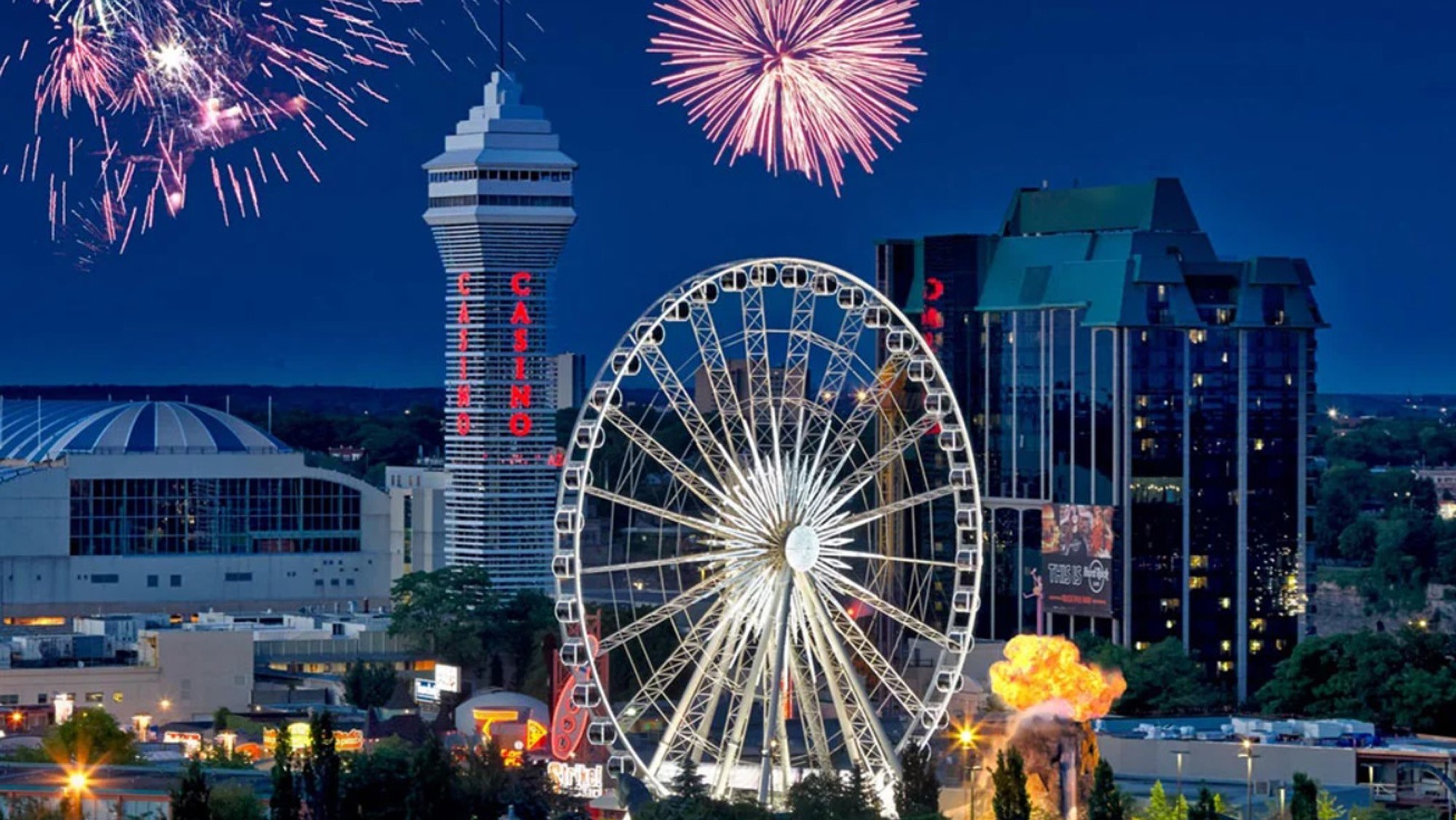 Colorful fireworks illuminate the city skyline, with a ferris wheel in the foreground, casino building in background.