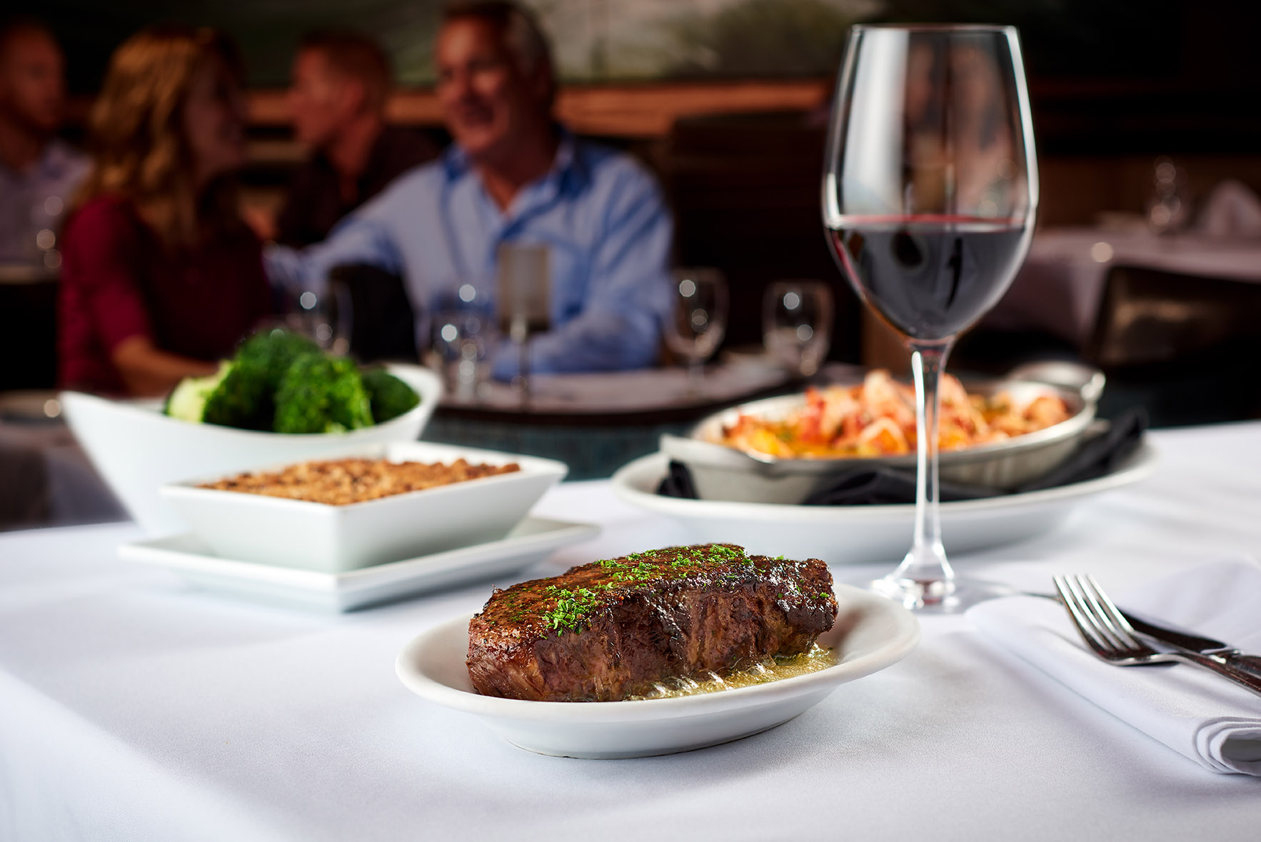 A table in a restaurant with a plate of steak, a bowl of broccoli, a seafood dish, and a glass of red wine, with people conversing in the background.