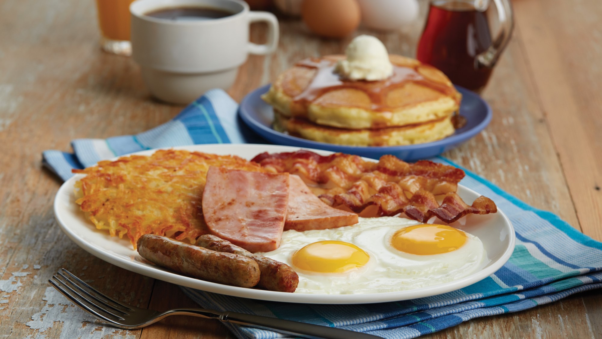 Hearty breakfast plate with bacon, eggs, sausage, ham, hash browns, and a side of pancakes topped with syrup and butter, accompanied by coffee and orange juice.