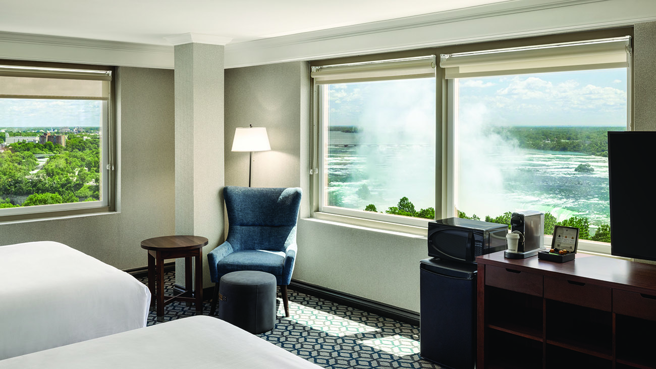 A hotel room with two queen beds with white linen, two large windows with a view of Niagara Falls, a spacious desk for work and a television.