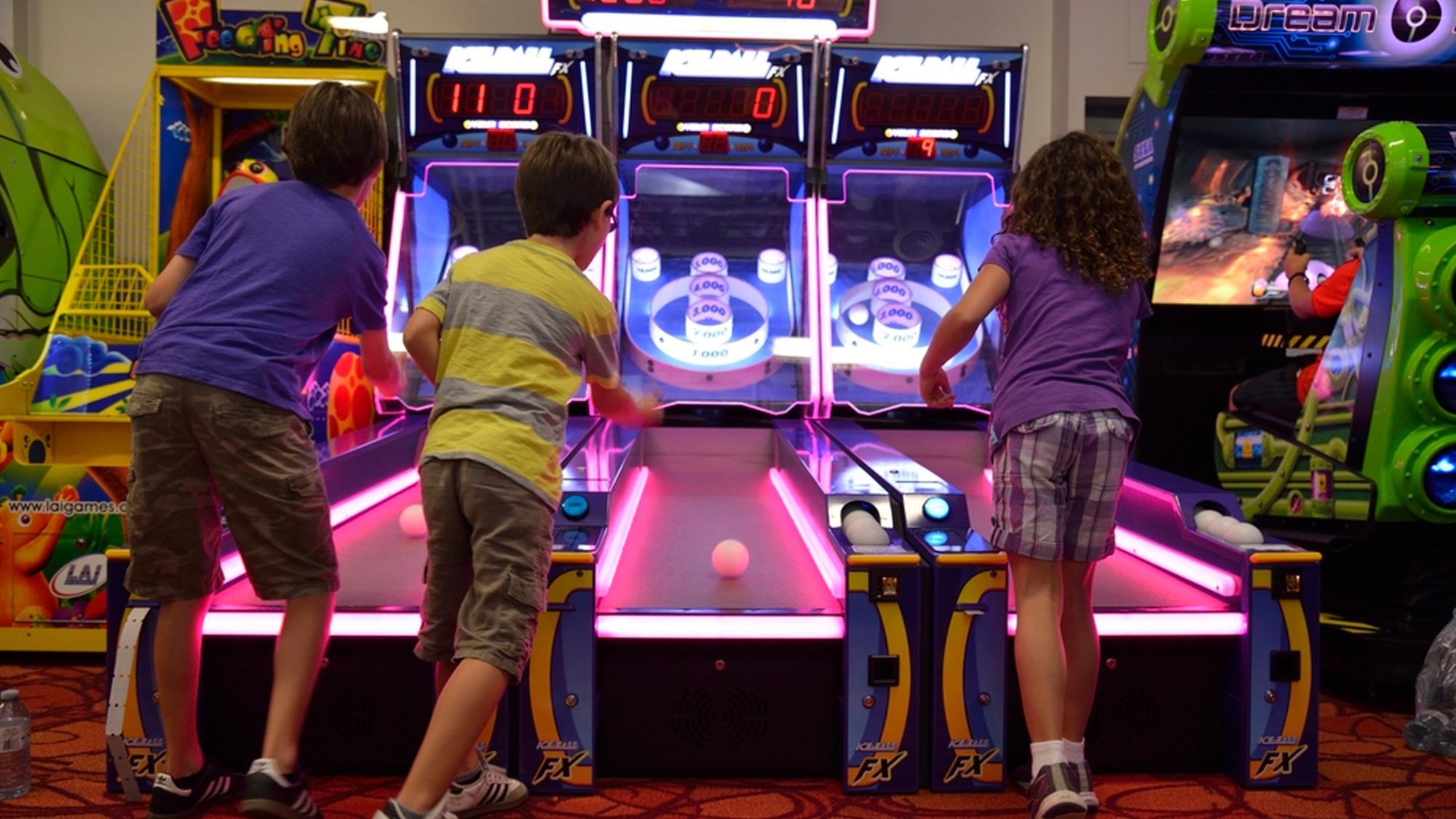 Three children playing skee-ball arcade games in a brightly lit game room.
