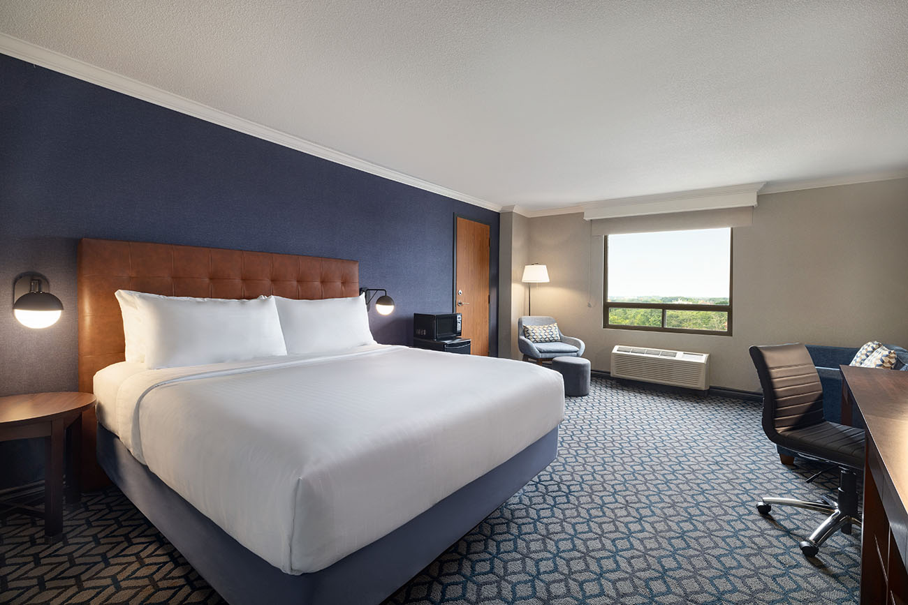 A hotel room with a king bed with white linen, a window offering view of city of Niagara Falls and a spacious desk for work or study purposes.