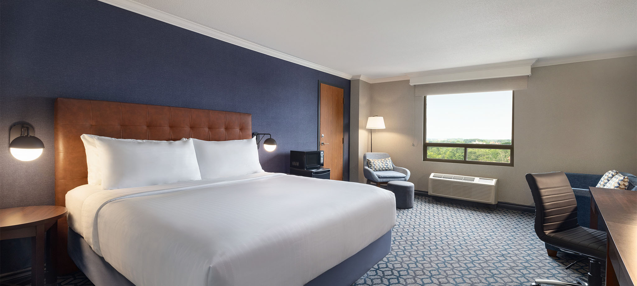 A hotel room with a king bed with white linen, a window offering view of city of Niagara Falls and a spacious desk for work or study purposes.