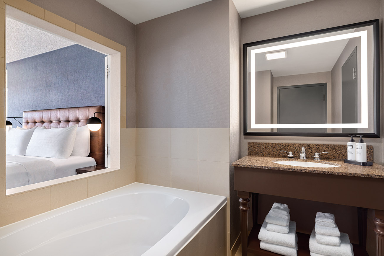 A bathroom scene displaying a relaxing whirlpool tub alongside window open to a bedroom with a king bed.