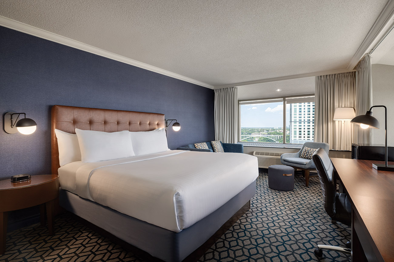 A hotel room with a king bed with white linen, a window offering view of city of Niagara Falls and a spacious desk for work or study purposes.