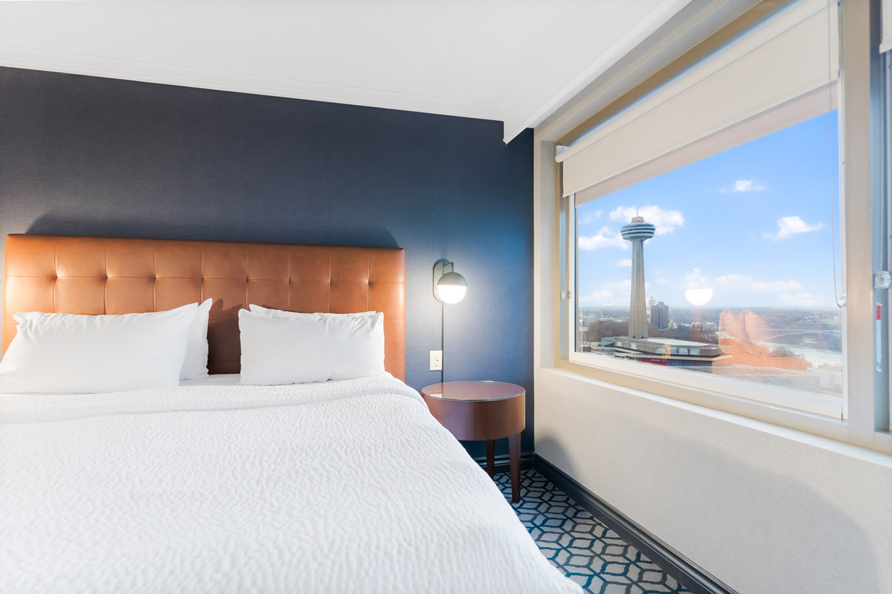 A hotel room with a king bed with white linen, a window offering view of city of Niagara Falls and Skylon Tower.