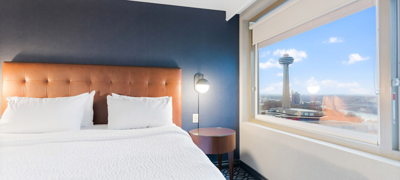 A hotel room with a bed with white linen, a window offering view of city of Niagara Falls and Skylon Tower.