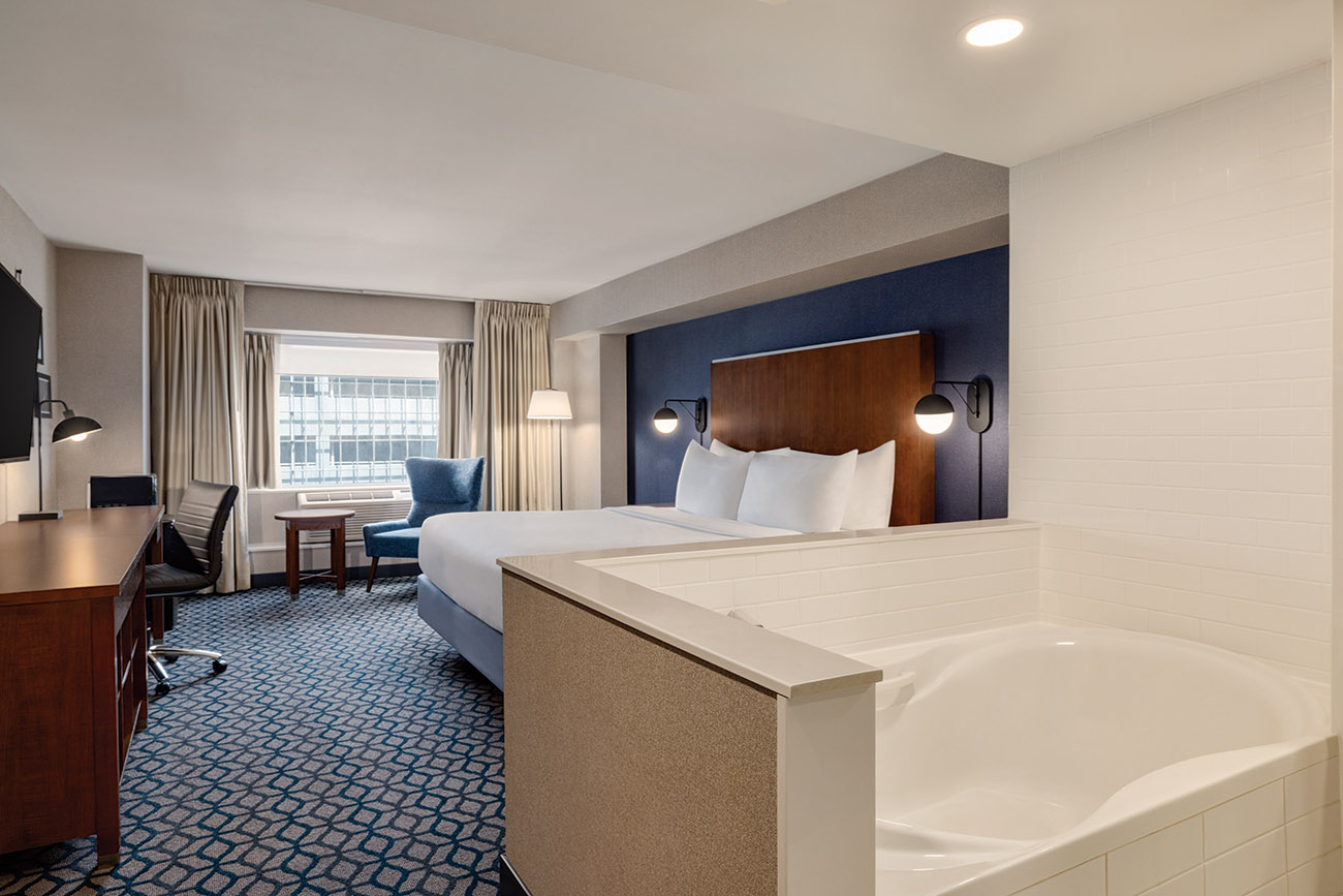 A hotel room with a whirlpool tub open to a bedroom with king bed with white linen, a window offering view of city of Niagara Falls.