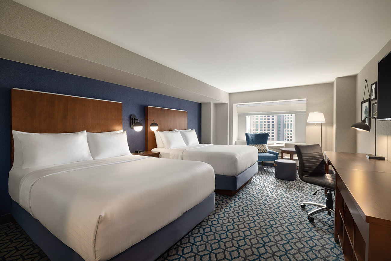 A hotel room with two queen beds with white linen in front of large window with view of the city. 