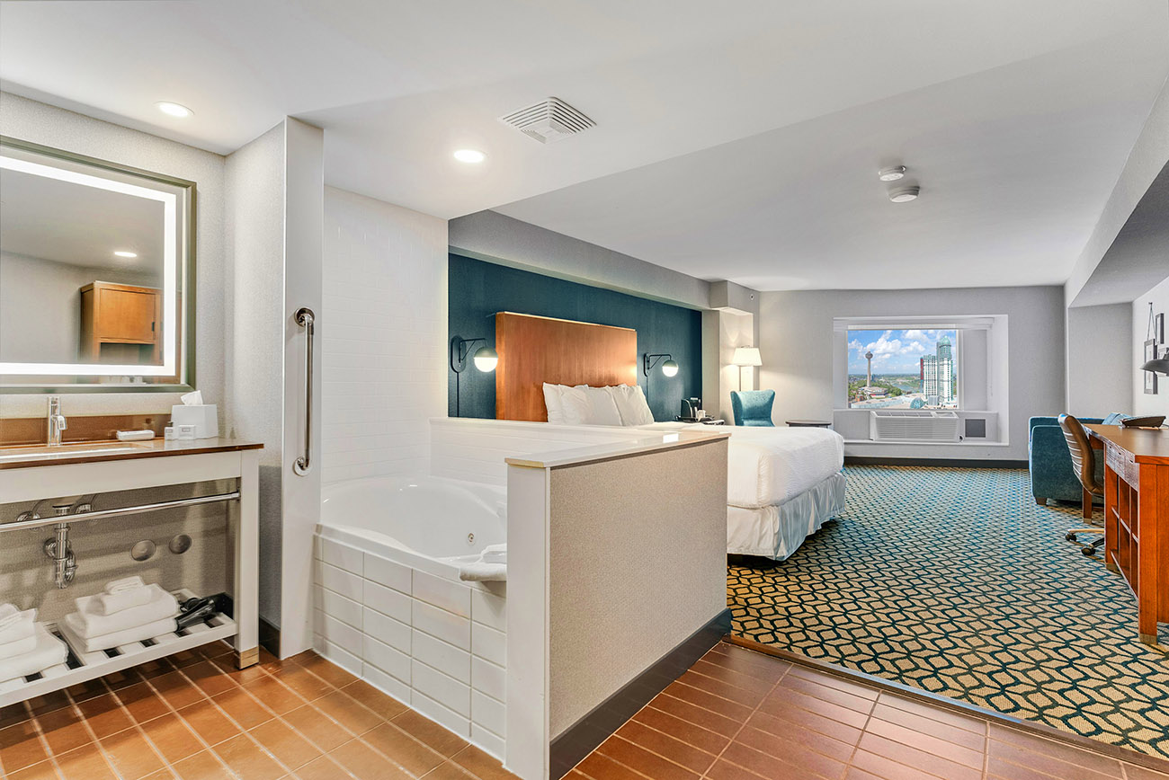 A hotel room with a whirlpool tub open to a bedroom with king bed with white linen,  a window offering view of city of Niagara Falls and Skylon Tower.