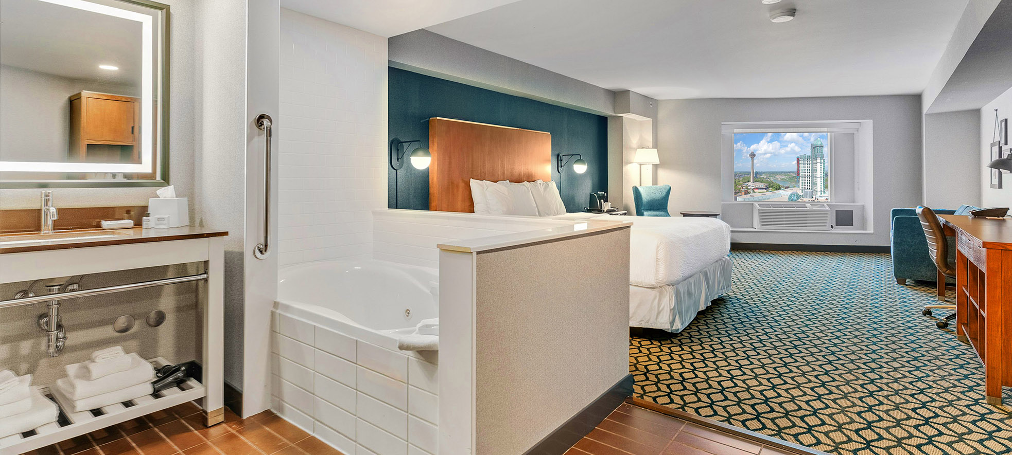 A hotel room with a whirlpool tub open to a bedroom with king bed with white linen, a window offering view of city of Niagara Falls and Skylon Tower.