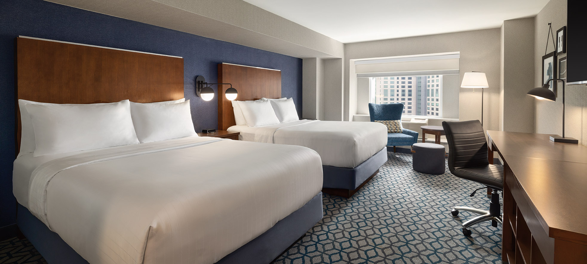 A hotel room with two queen beds with white linen, a window offering view of city of Niagara Falls and a spacious desk for work or study purposes.