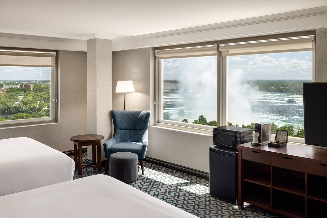 A hotel room featuring two queen beds, a television, and large windows allowing viewing Niagara Falls.