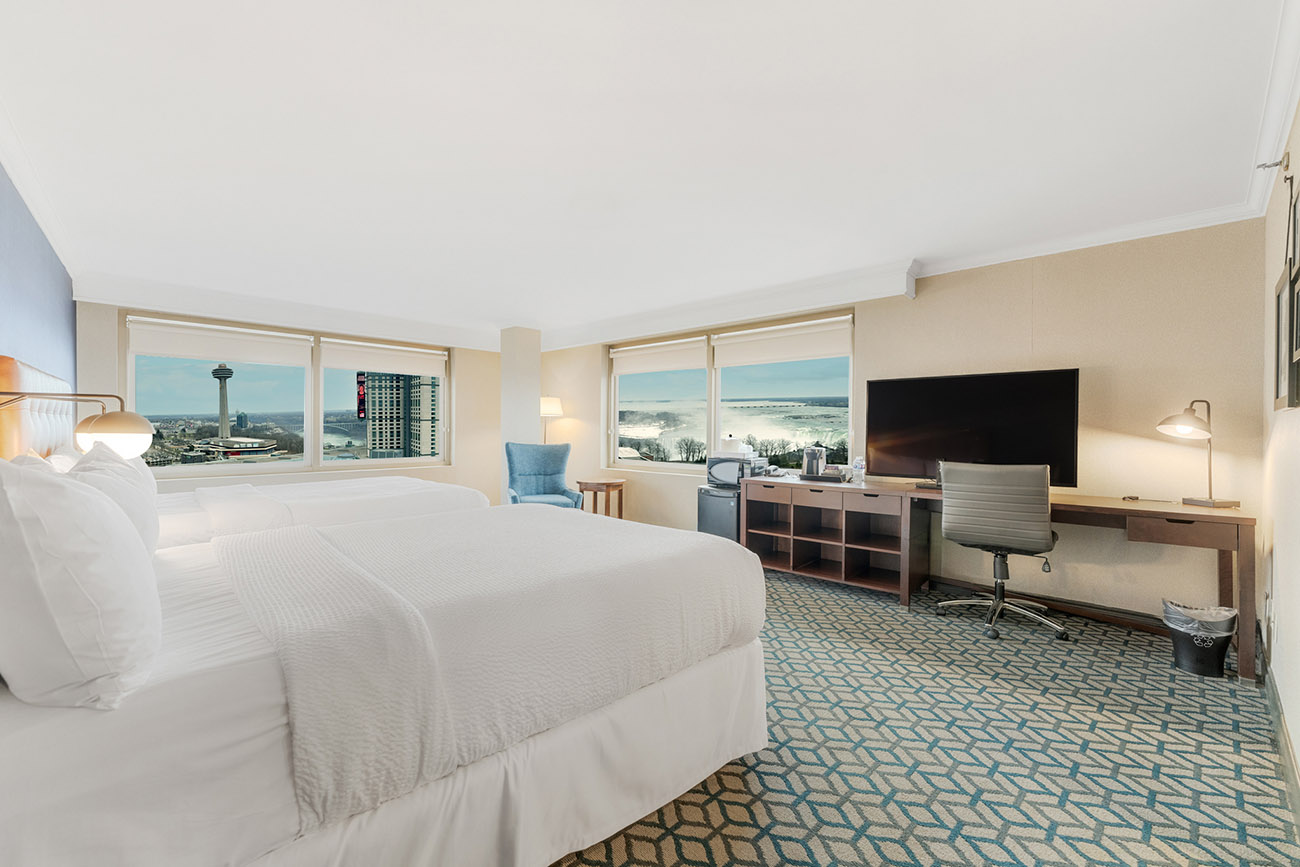 A hotel room with two queen beds with white linen, two large windows with a view of Niagara Falls and a spacious desk for work and a television.