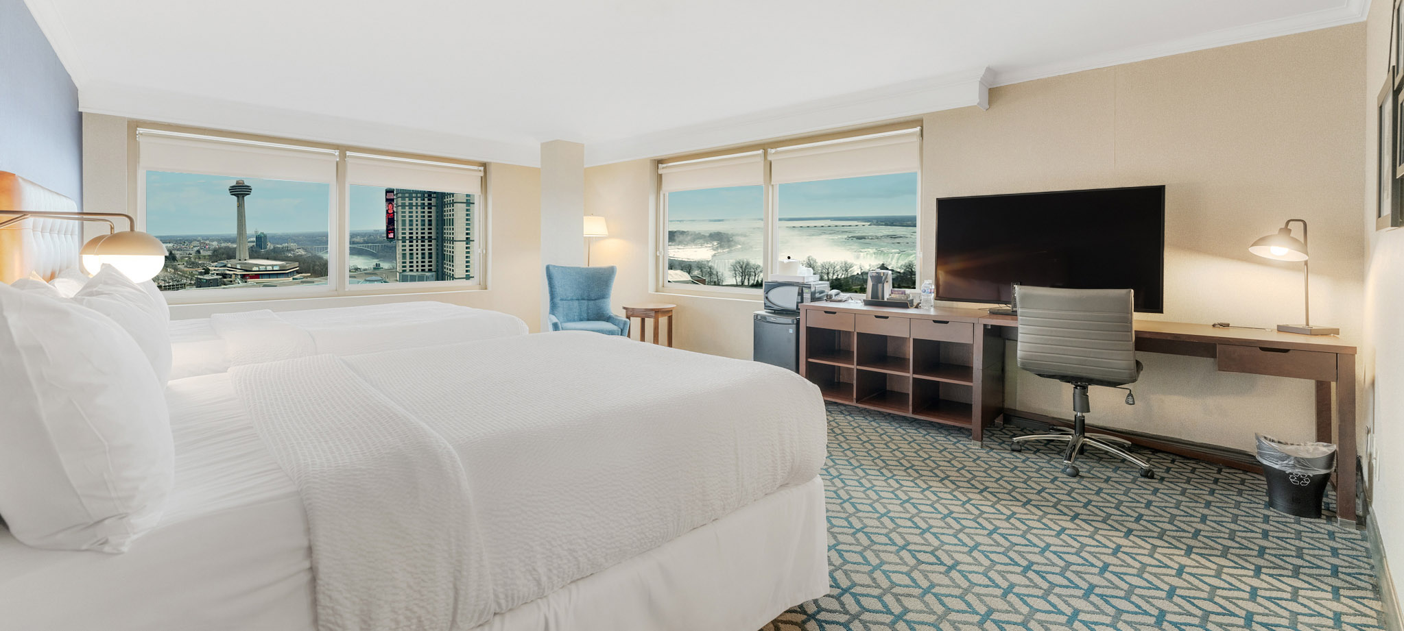 A hotel room with two queen beds with white linen, two large windows with a view of Niagara Falls and a spacious desk for work and a television.