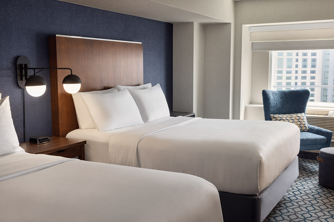 A hotel room with two queen beds with white linen in front of large window with view of the city. 