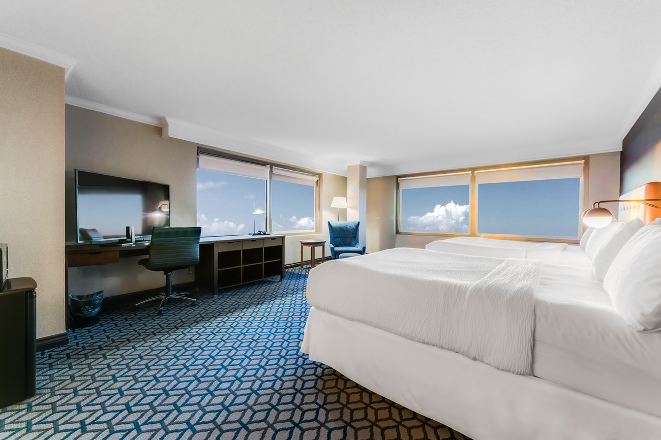 A hotel room with two queen beds with white linen, two windows offering view of city of Niagara Falls and a spacious desk for work and television mounted to the wall.