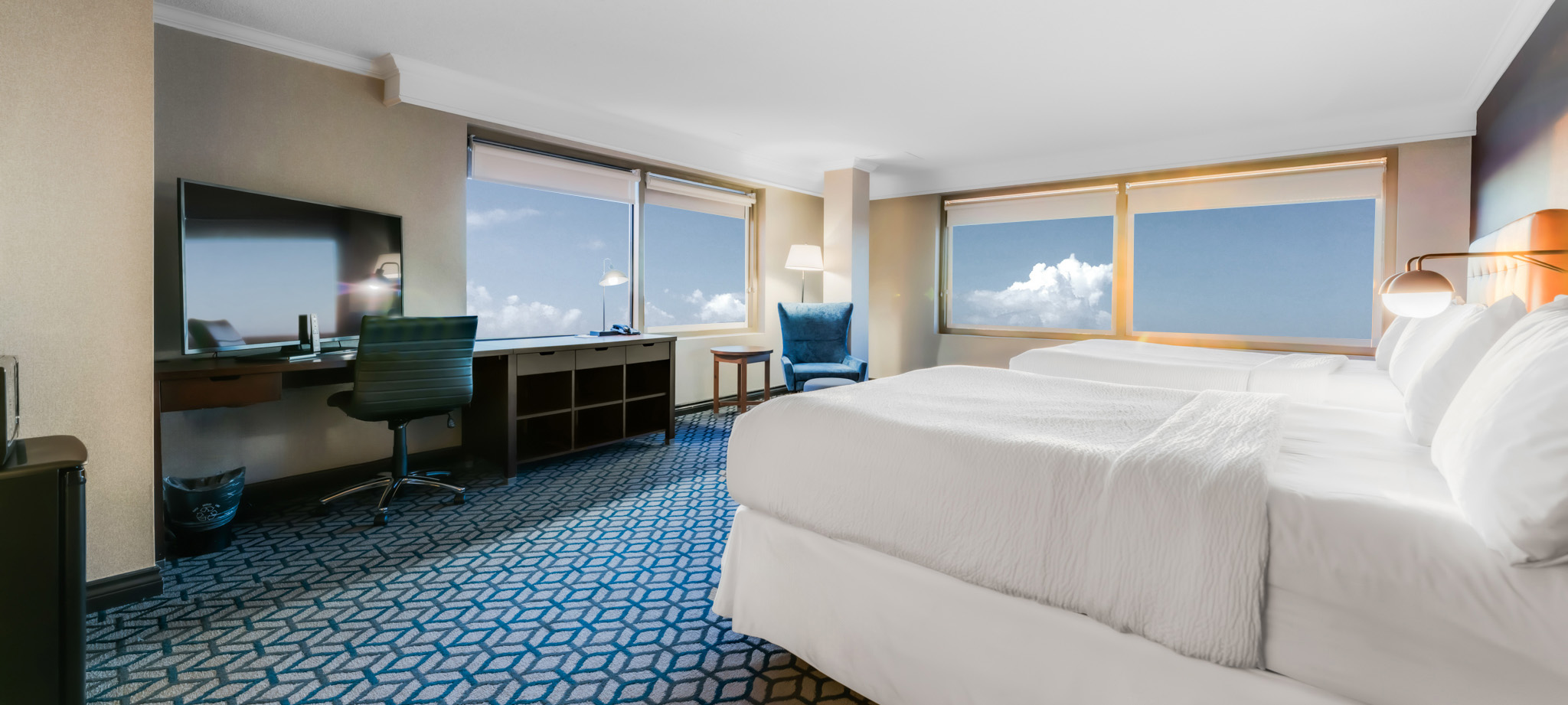 A hotel room with two queen beds with white linen, two windows offering view of city of Niagara Falls and a spacious desk for work and television mounted to the wall.